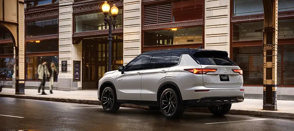 2025 Mitsubishi Outlander PHEV parked on a city street, rear-side view showcasing its modern design and black alloy wheels.